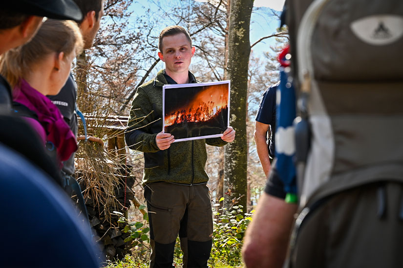 Joshua Huber hält während einer Feuerwehrschulung zum Thema Waldbrände ein Foto eines Waldbrands hoch und zeigt es einer Gruppe Teilnehmender im Freien.